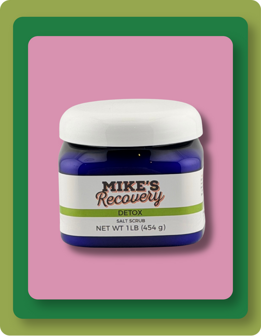 A jar of Mike's Recovery Detox Salt Scrub, featuring a dark blue container, a white lid, and a white label with green and grey  text, set against a pink background with green and orange borders.