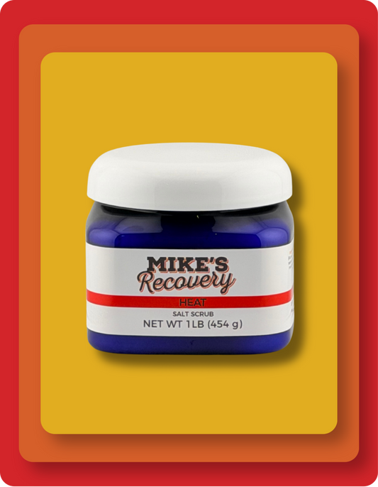 A jar of Mike's Recovery Heat Salt scrub  with a blue container, white lid, and white label with red and black text, set against a yellow and orange graphic background.
