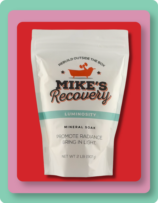 A stand-up pouch of Mike's Recovery Luminosity Mineral Soak, featuring a white bag with orange and black lettering and a teal band, set against a red background with a pink and mint border.