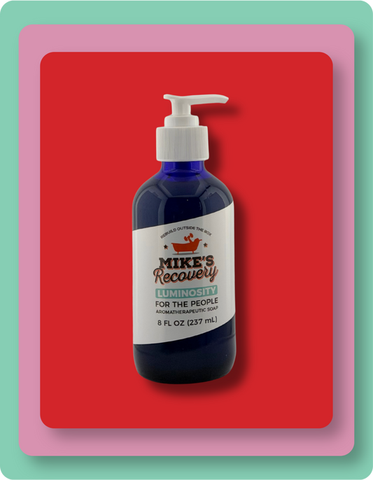 A dark blue pump bottle of Mike's Recovery Luminosity hand and body soap set against a bold red background with a pink and mint green layered border.