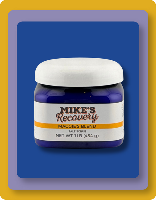 A jar of Mike's Recovery Salt Scrub, Maggie's Blend, featuring a white cap and a white label with orange and brown text, set against a blue background with a yellow border.