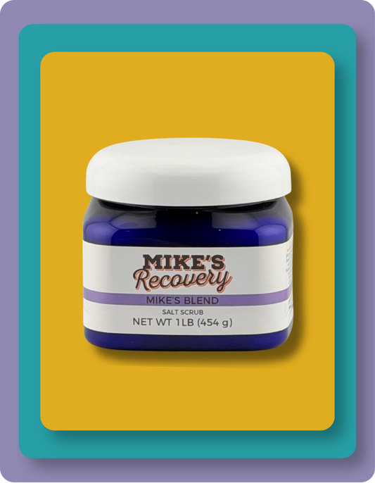 A jar of Mike's Recovery Salt Scrub, 'Mike's Blend', in a dark blue jar with a white lid and label, set against a yellow background with a teal and purple border.